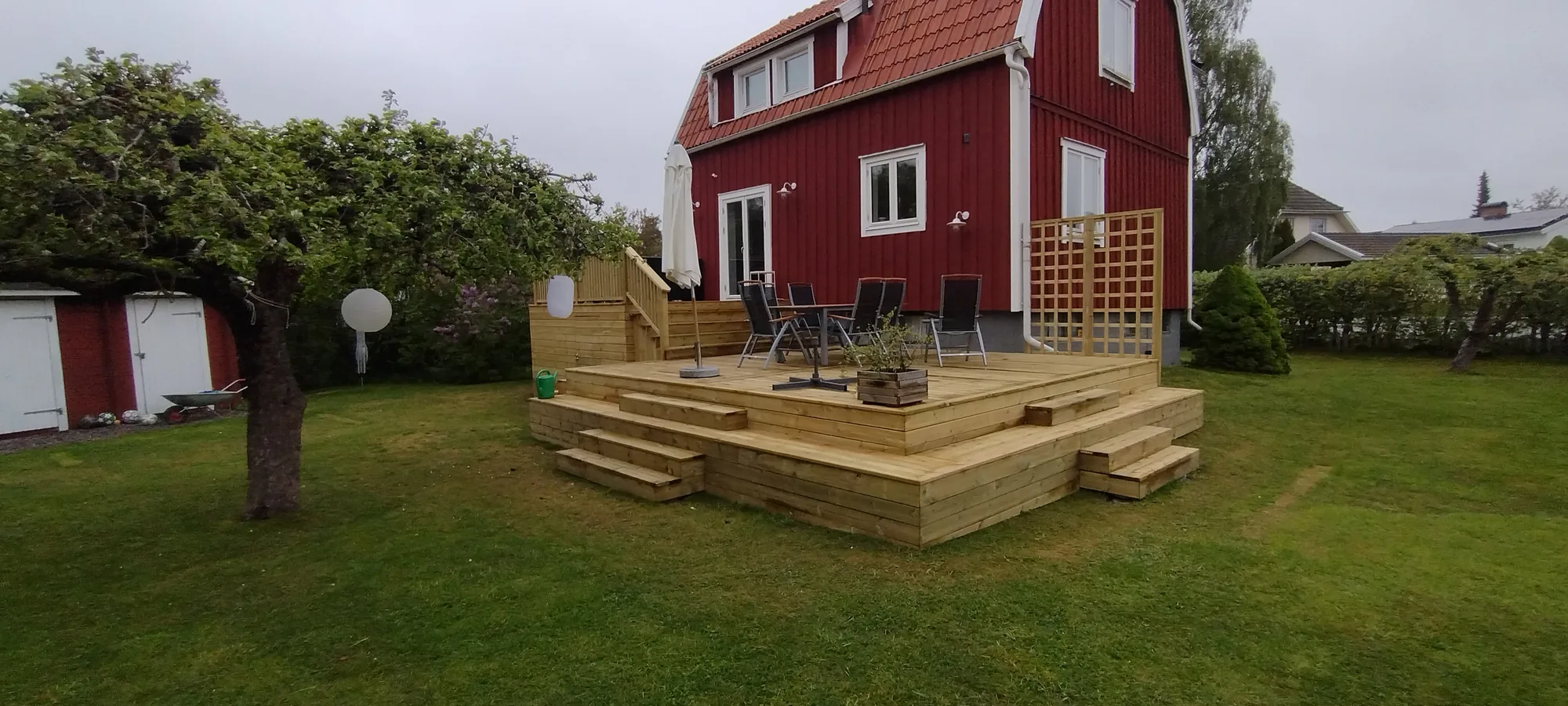 Nybyggd altan i tryckimpregnerat trä med sittsteg och spaljé framför ett falurött trähus